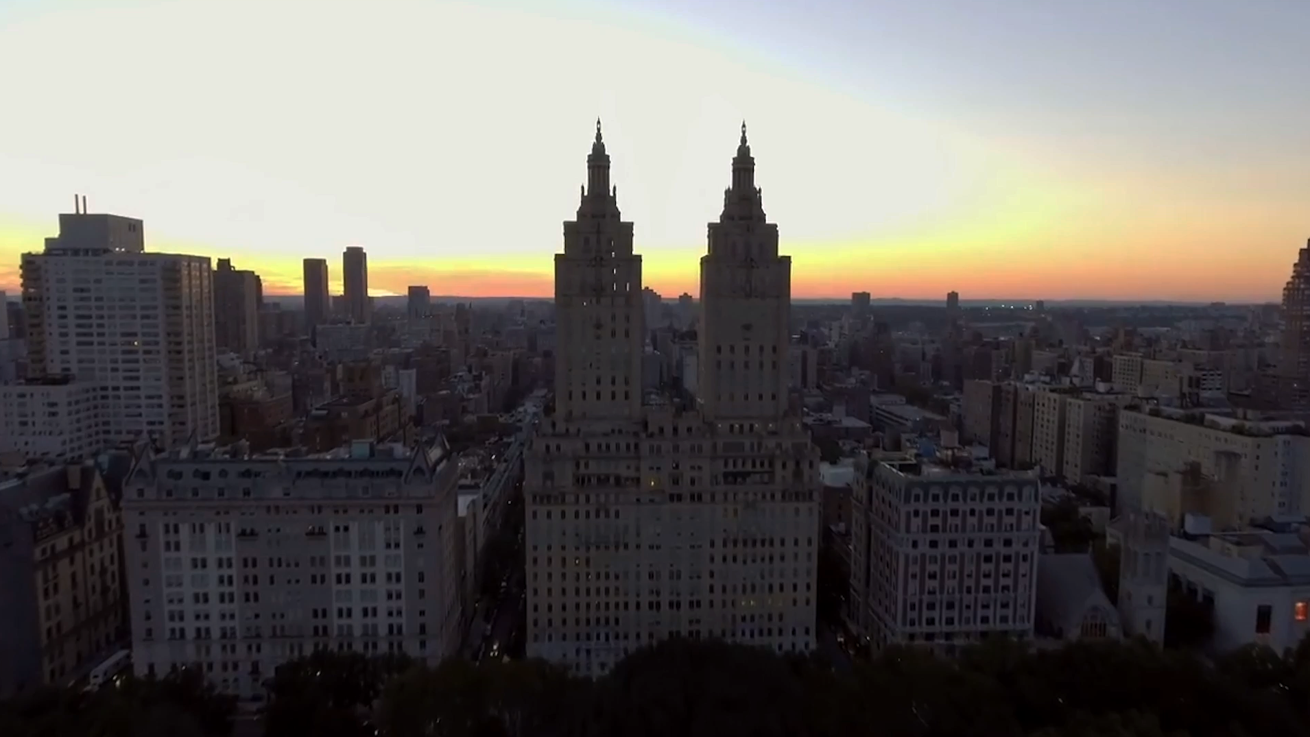 Aerial view of a cityscape during twilight with the silhouette of two prominent, ornate towers against a gradient sky transitioning from orange to blue. The surrounding buildings are in shadow, highlighting the towers as focal points.